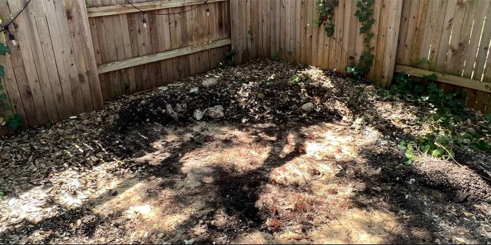 After: the same fenced corner with the hot tub and its pad gone, leaving raked bare earth and mulch running clean into both fence lines.