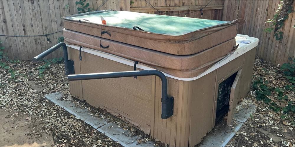 Before: a disused beige hot tub with a torn cover sitting on a concrete pad in a leaf-strewn corner between two wooden fences, its pipework hanging loose down the side.