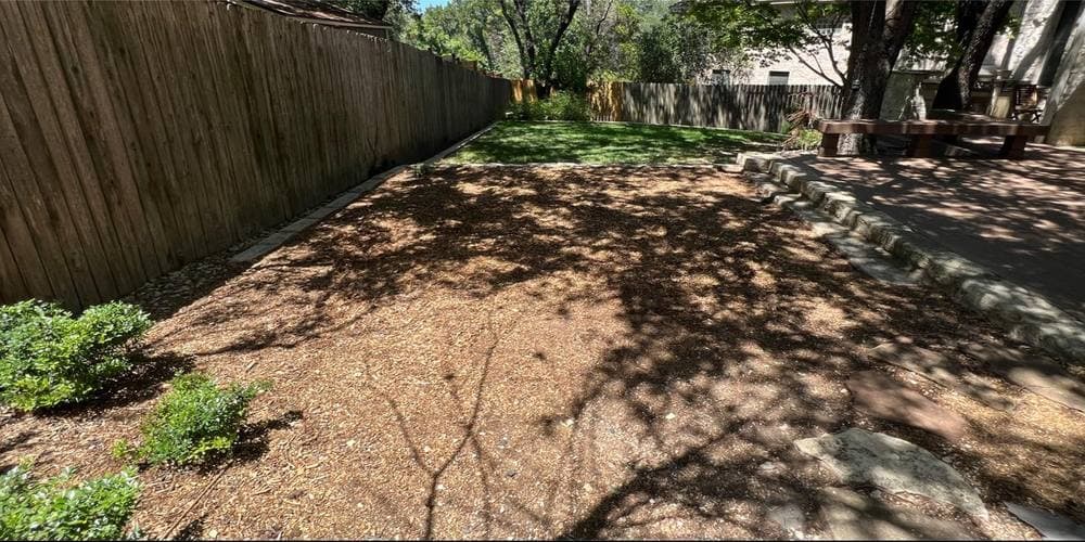 After: the same fence line with the playscape gone, an open run of level mulch and grass stretching back to a low stone border.