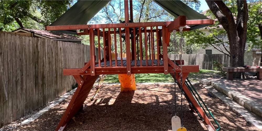 Before: a large red-stained wooden playscape with a climbing platform, a canopy, a slide and swings, standing on mulch tight against a wooden fence.