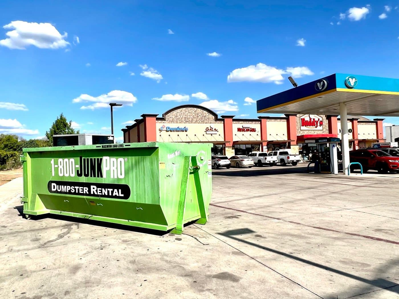 A green 1-800-JUNKPRO roll-off dumpster parked in a sunny strip-mall lot with a gas station and storefronts behind it.