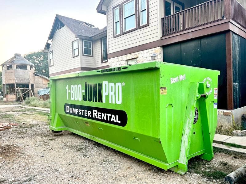 An empty green roll-off container standing on a bare gravel lot beside a half-finished two-storey house, its side panel reading 1-800-JUNKPRO Dumpster Rental.
