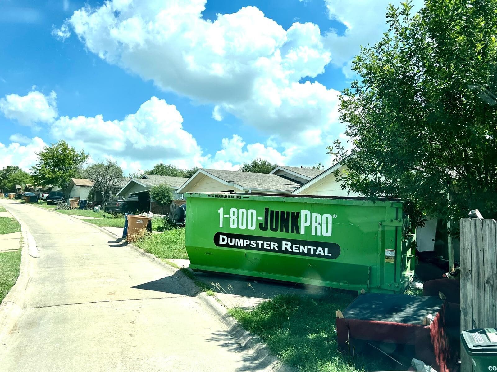 A green roll-off container at the curb of a residential street with a couch and other bulk items staged beside it for pickup.