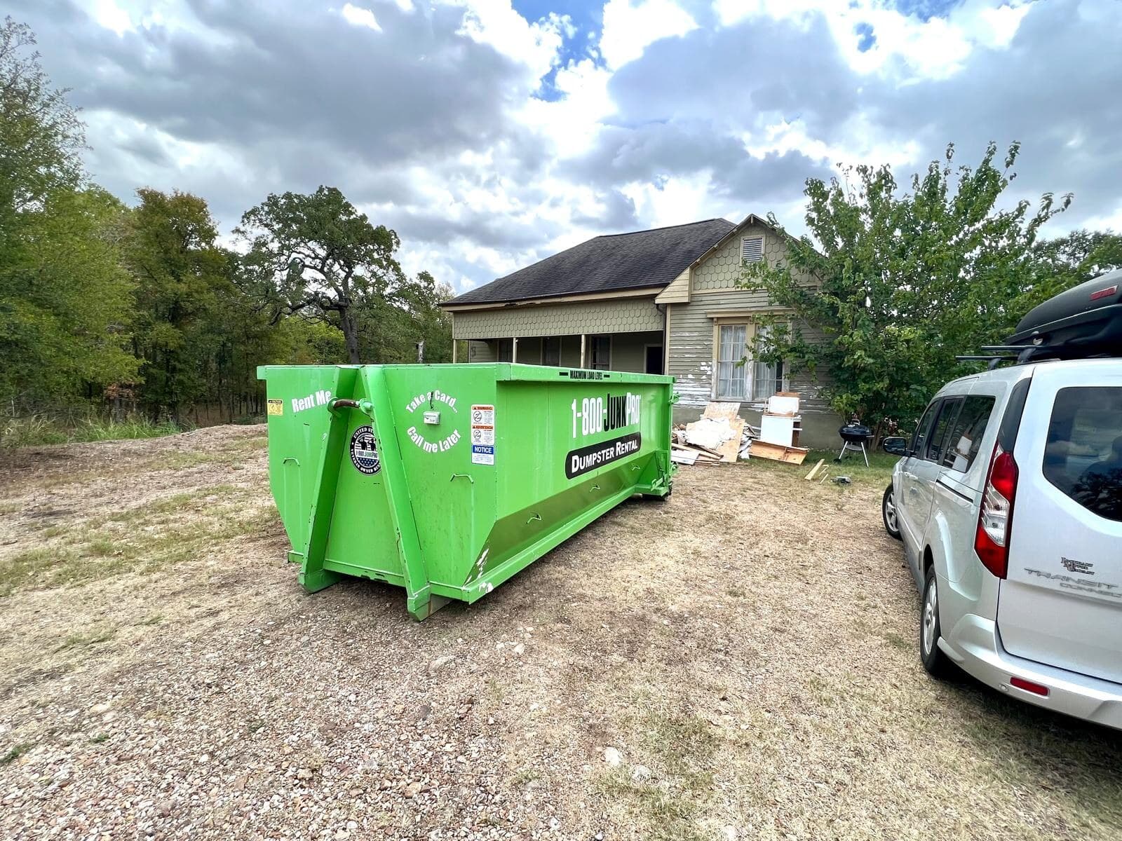 An empty green roll-off container on a gravel drive in front of an older wood-sided farmhouse, a van parked alongside.