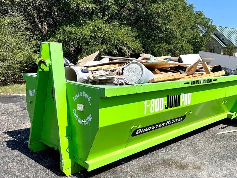 A green roll-off container loaded to the maximum load line with broken drywall, cut framing lumber, buckets and offcuts from a remodel, parked on asphalt under live oaks.