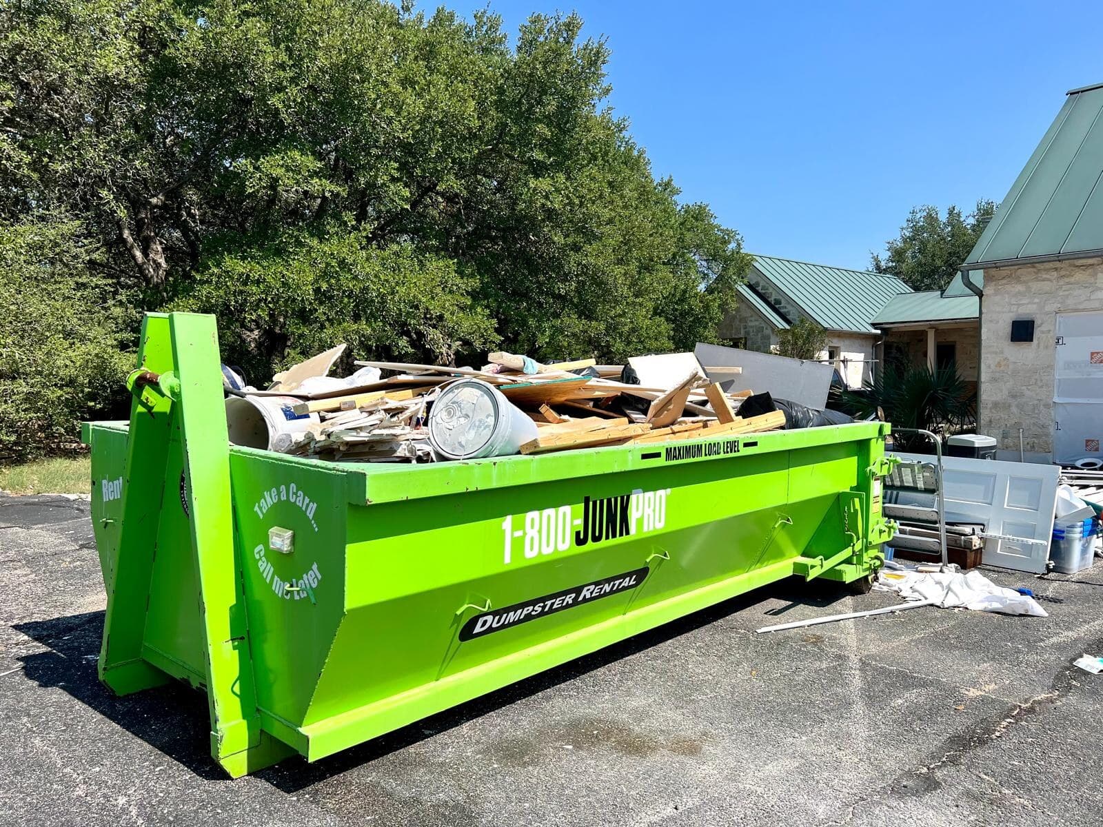 A green roll-off container loaded with lumber and construction debris, parked beside a stone building with a green metal roof.