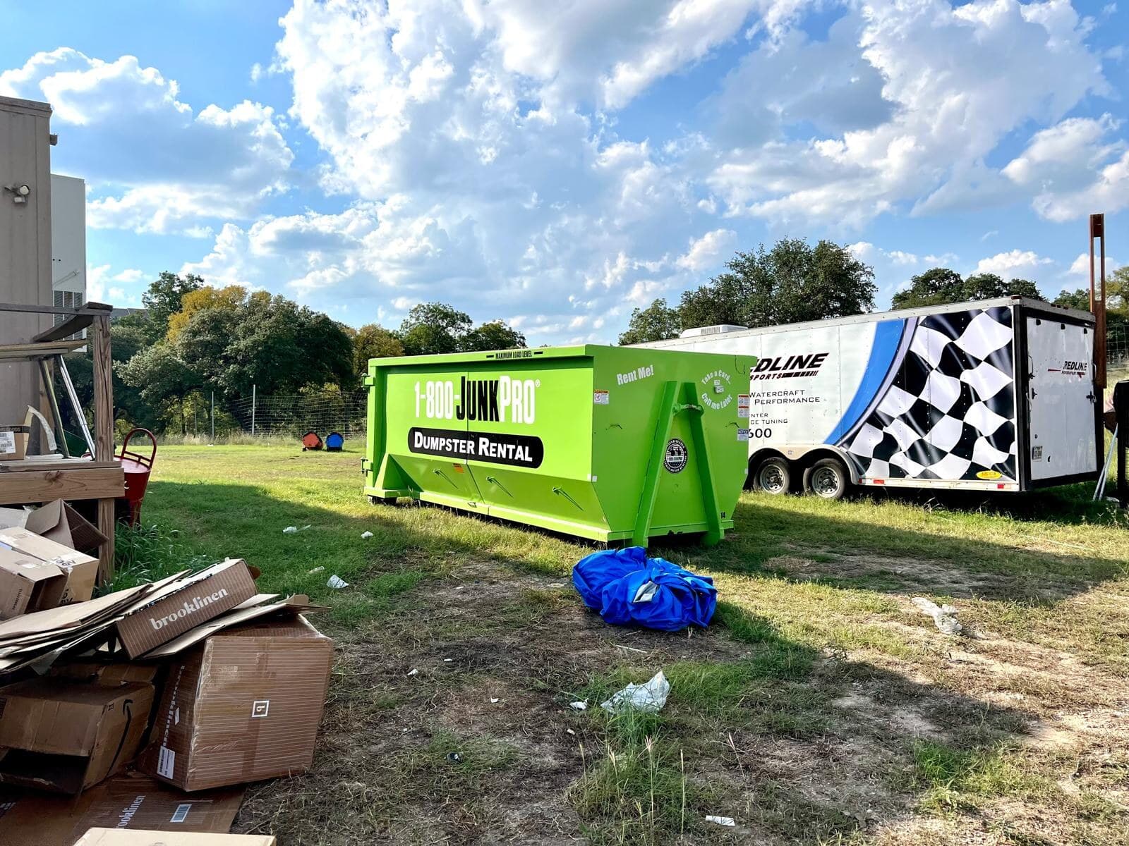 An empty green roll-off container on open grass with an equipment trailer parked nearby.