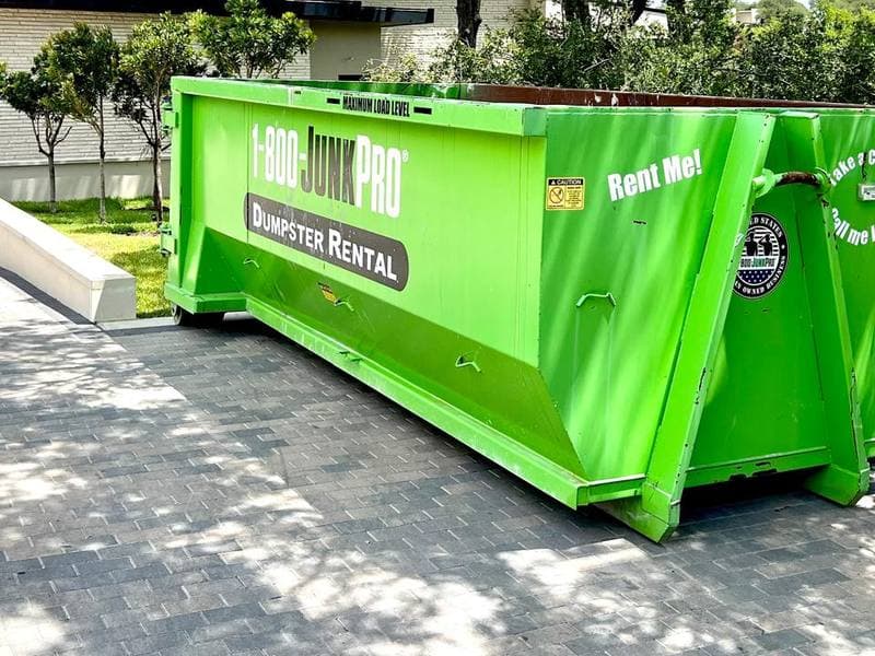 A long green roll-off container set down lengthways on a grey paver driveway, resting square on the pavers with a clipped row of young trees and a limestone wall behind it.