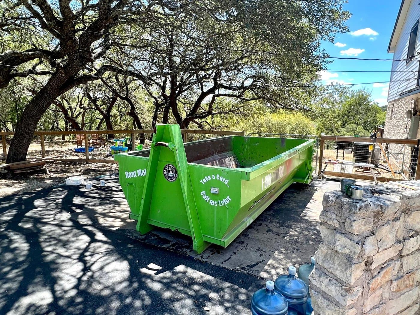 A long green roll-off container set on a shaded driveway next to a limestone wall, live oaks arching overhead.