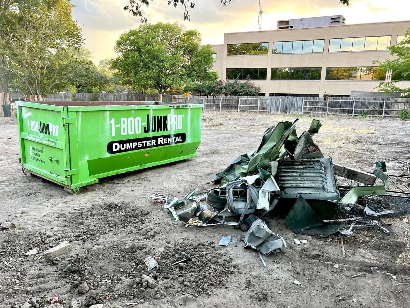 A green roll-off container on a bare dirt lot beside a crushed pile of scrap sheet metal, an office building in the background at sunset.