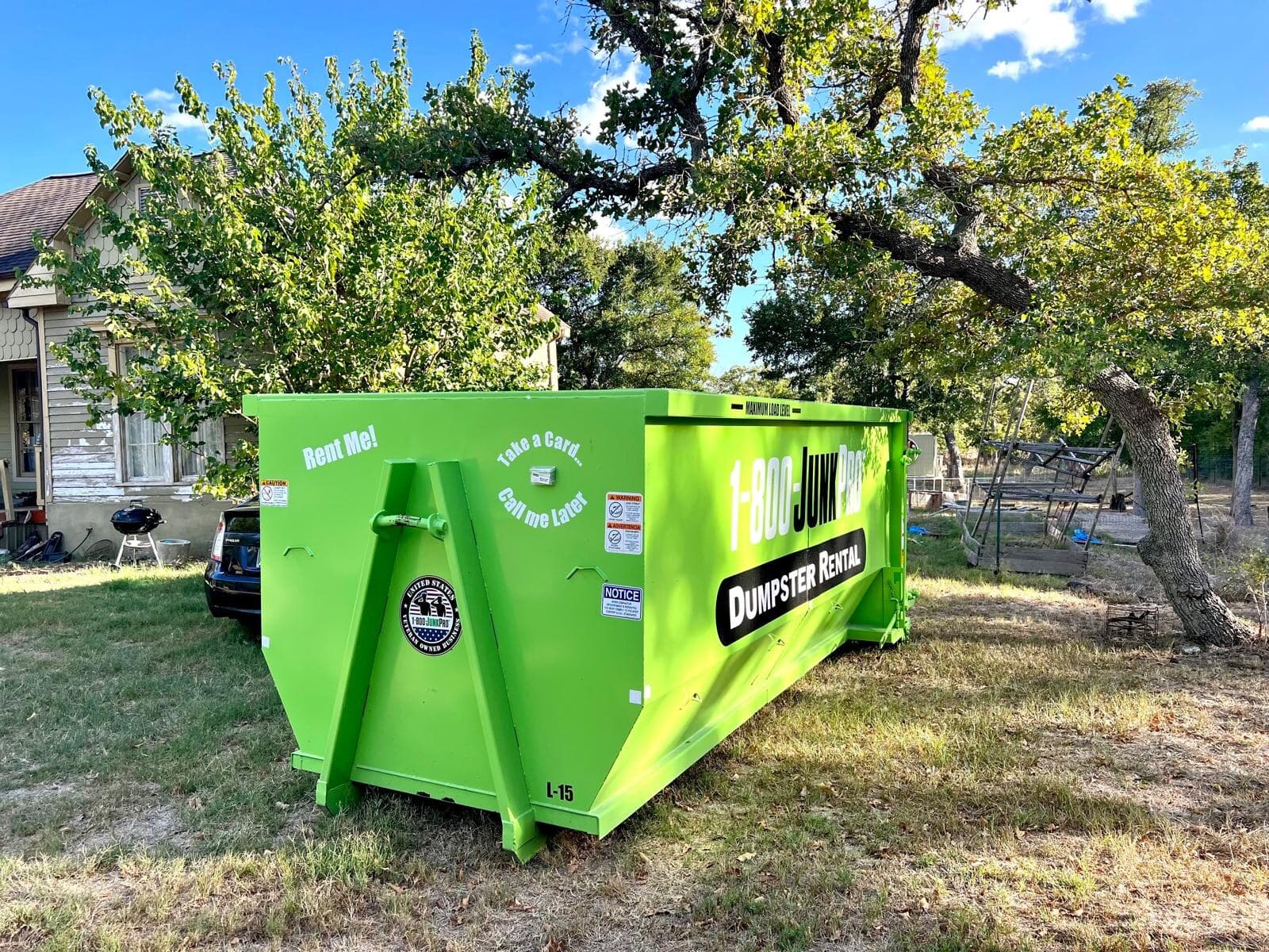 A loaded green 1-800-JUNKPRO roll-off container in a driveway in front of a tan brick house, an SUV parked beside it under a large oak tree.