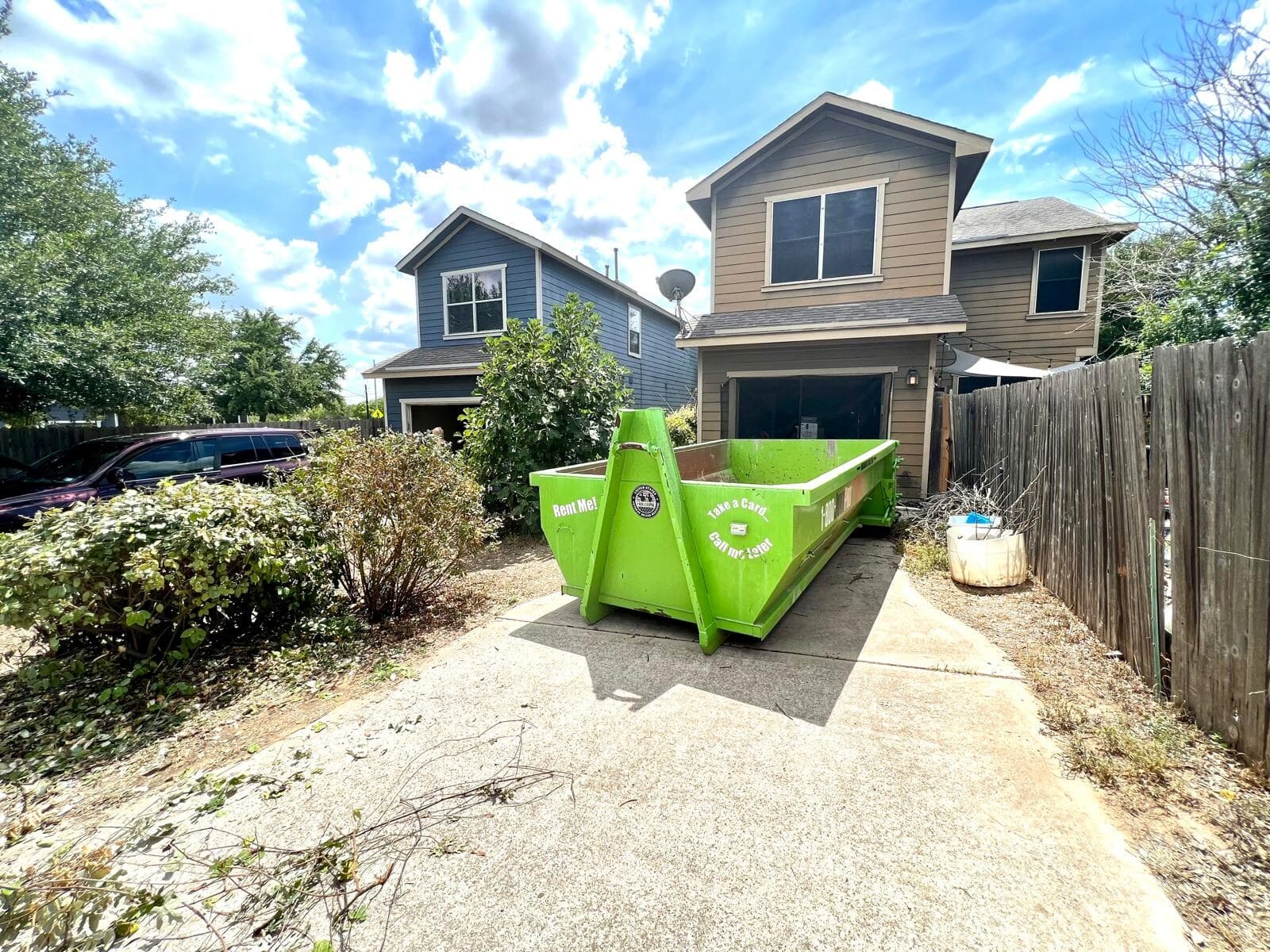 An empty green roll-off container in a driveway between two-story townhomes, an SUV parked in the neighboring drive.