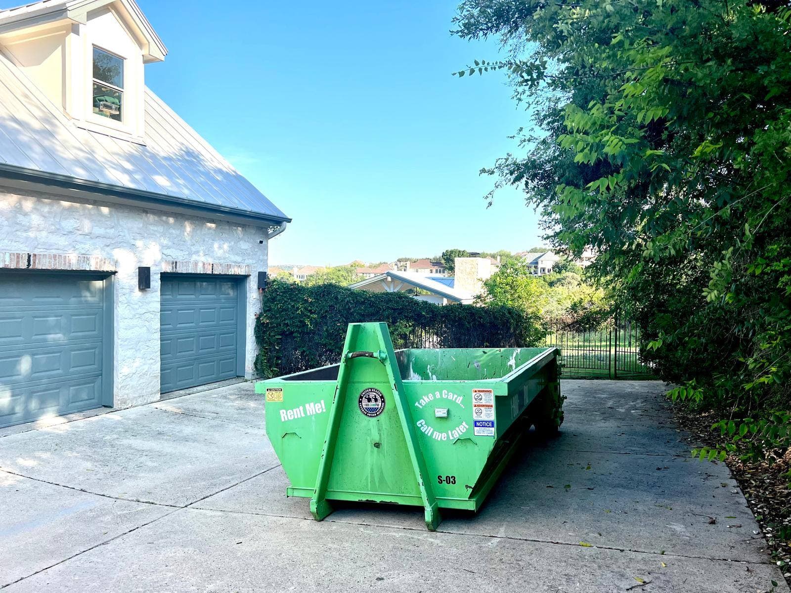 An empty green roll-off container standing on the concrete driveway of a stone Austin house, parked clear of the garage doors with room left to get a car past it.